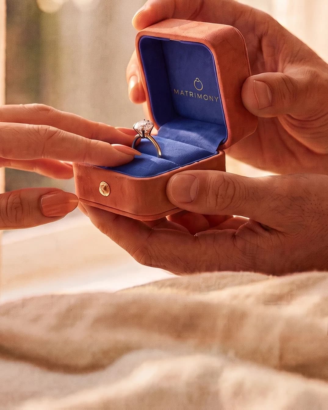 Él abre el estuche Matrimony de gamuza coral y ella acerca la mano al anillo de compromiso